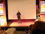 Full view of stage with Douglas Adams standing in the middle of a carpet with no props or lectern