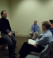 Joel leaning against a table facing people on chairs with Don in the background listening