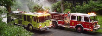 Two fire trucks and smoke on a narrow street with lots of trees