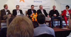 Six people sitting up on a low podium in front of people sitting in chairs